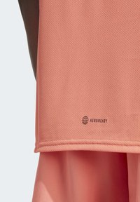 Camiseta deportiva de malla en color durazno con branding AEROREADY que absorbe la humedad en el dobladillo. La textura es transpirable con un forro interno suave.