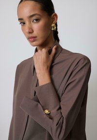 Blusa marrón de cuello con una textura suave, que presenta un detalle de botón en la manga. El modelo lleva el cabello recogido y un maquillaje natural.