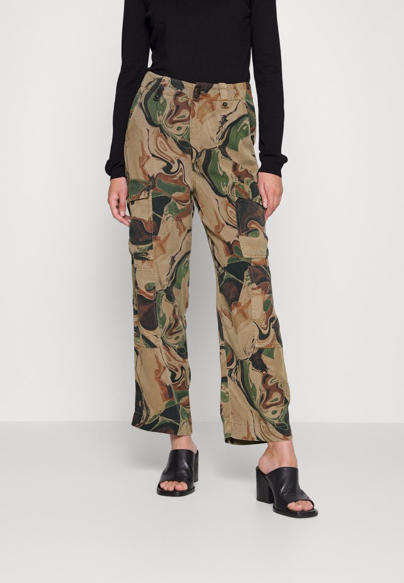 Calças cargo camufladas em tons de bege, verde e castanho, com um ajuste folgado, múltiplos bolsos e detalhes de costura visíveis, combinadas com mules pretos de biqueira aberta.