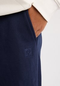 Navy blue sweatpants made of soft fabric, featuring a small embroidered logo on the thigh and side pockets.