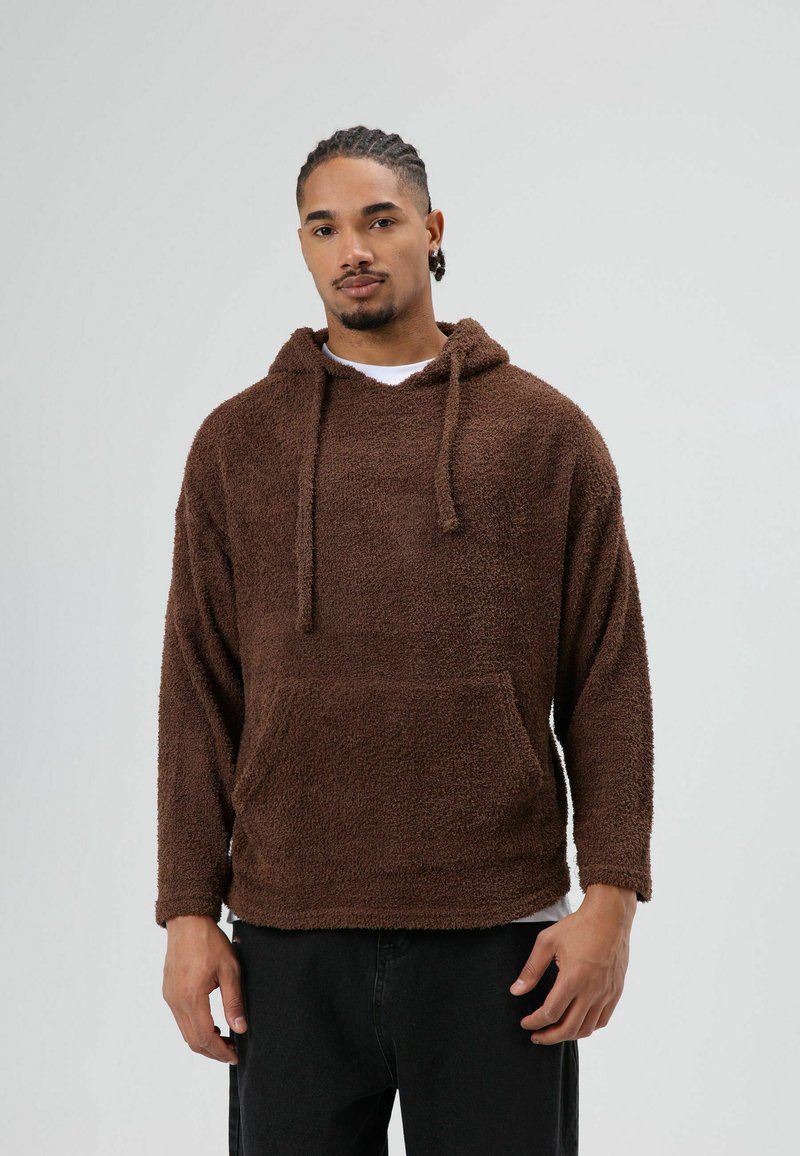Sweat à capuche marron texturé avec capuche à cordon de serrage et poche avant. Le tissu semble doux et moelleux, avec des manches longues et une coupe décontractée.