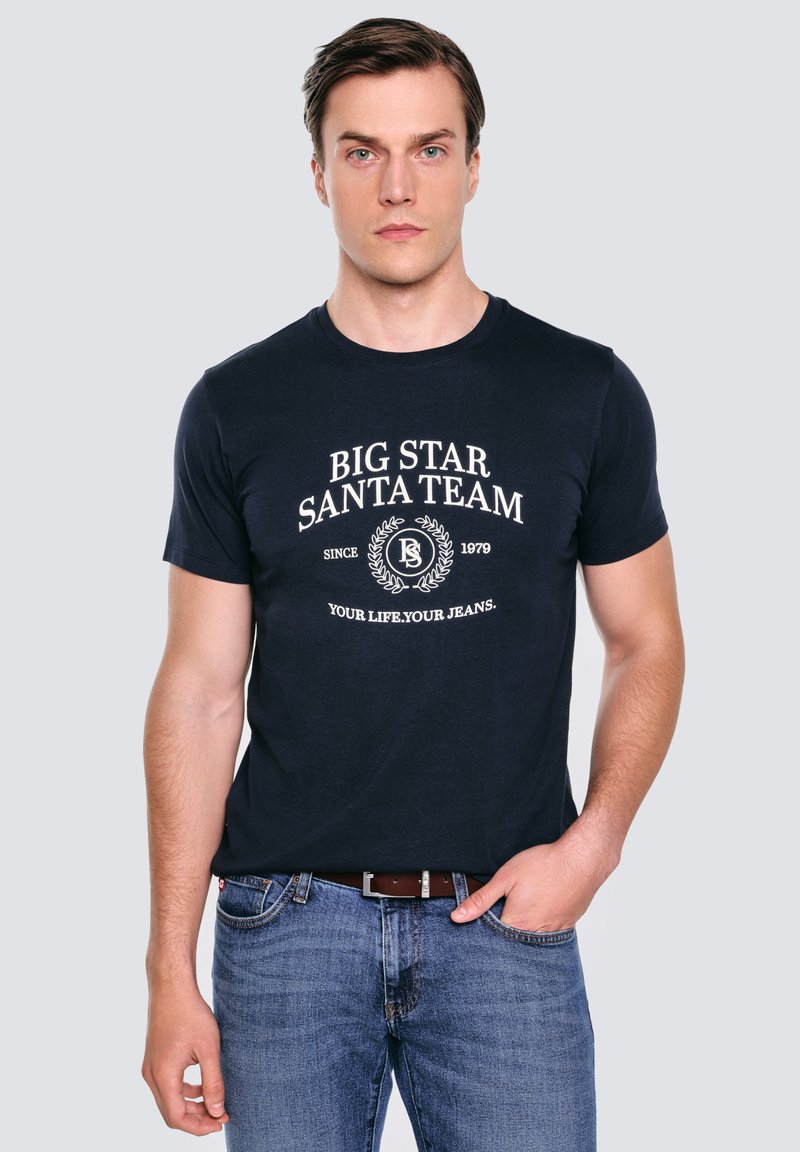 Námořnická modrá bavlněná tričko s bílou kruhovou grafikou a textem "BIG STAR SANTA TEAM" a designem vavřínového věnce. Nosič má na sobě modré džíny.