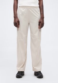 Άτομο που φοράει άνετο μπεζ παντελόνι και μαύρα παπούτσια πεζοπορίας, στέκεται μπροστά σε λευκό, απλό φόντο.