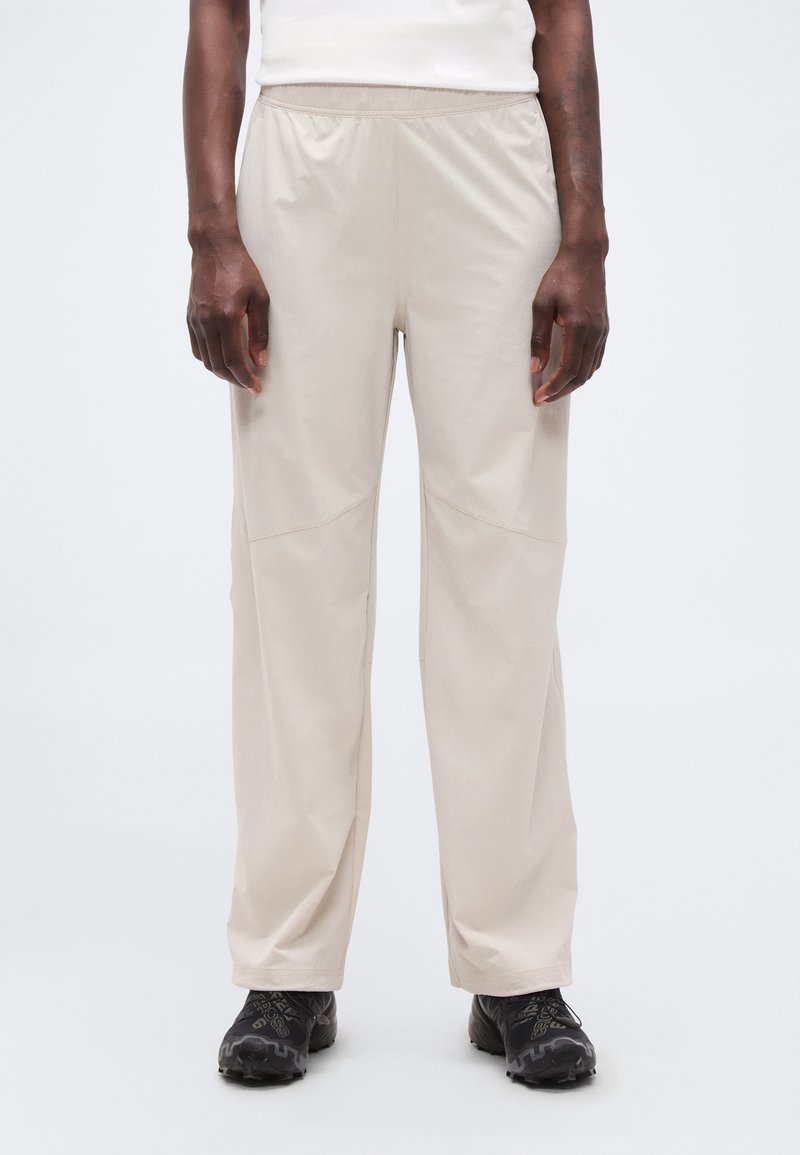 Άτομο που φοράει άνετο μπεζ παντελόνι και μαύρα παπούτσια πεζοπορίας, στέκεται μπροστά σε λευκό, απλό φόντο.