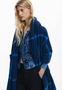 Châle à carreaux bleu marine avec texture et accents à franges drapé sur un haut à motifs floraux bleus. Le mannequin se tient avec les mains dans les poches.