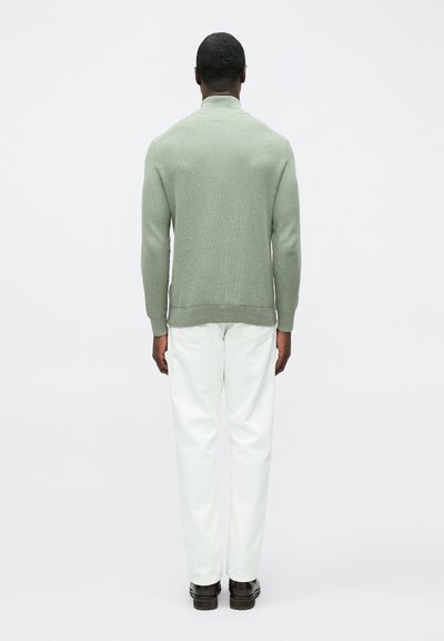 Világoszöld pulóver magas gallérral, kötött textúrával és bordázott mandzsettákkal, fehér nadrággal és fekete cipővel párosítva, hátulnézetben.