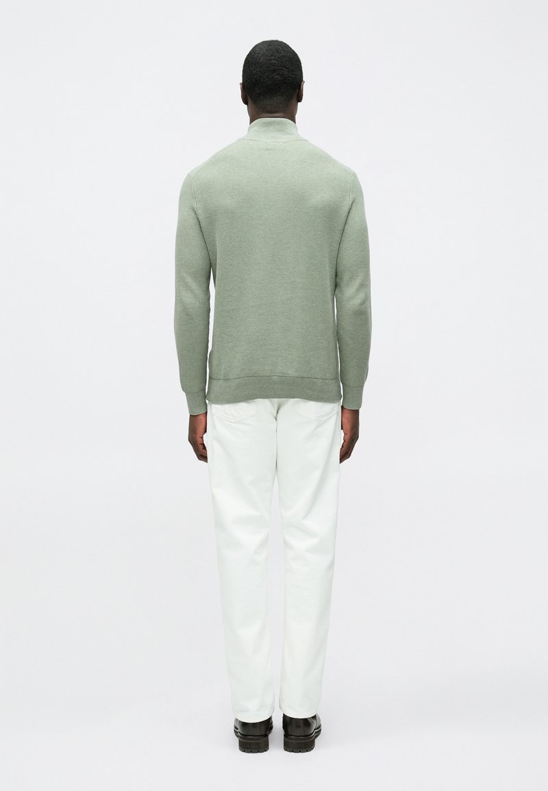 Maglione verde chiaro con collo alto, tessuto lavorato a maglia e polsini a coste, abbinato a pantaloni bianchi e scarpe nere, visto da dietro.