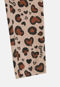 Leggings i en lys beige stof med et brunt og sort plettet mønster, inklusiv hjerter; indeholder mærkenavnet "Guess" i sort.