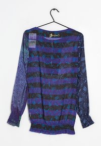 Blusa morada transparente con mangas largas, que presenta rayas negras y azules, patrones intrincados y una cintura fruncida. Colgada en una percha negra.