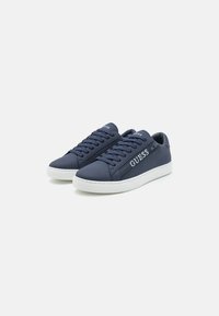 Tengerészkék sneaker, sima szintetikus anyagból készült, fehér gumitalppal, fűzős kivitelben, és a "GUESS" logóval az oldalon.