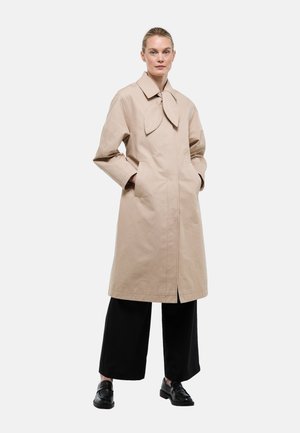 Femme portant un manteau beige mi-longues avec un col noué, un pantalon noir à jambes larges et des mocassins noirs, debout les mains dans les poches du manteau.
