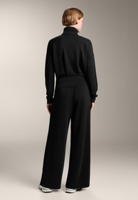 Černá souprava na volný čas obsahující pulovr s vysokým límcem a široké kalhoty. Měkká, jemná tkanina s volným střihem a decentní žebrovanou strukturou.