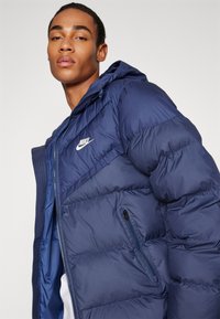 Casaco puffer azul-marinho com fecho de correr na frente, capuz ajustável e logótipo branco. Apresenta costura acolchoada e bolso lateral com fecho.