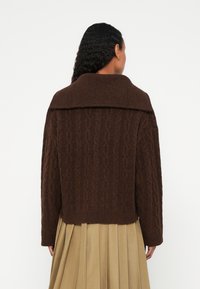 Žena s dlhými kučeravými vlasmi, oblečená v hnedom sveteri s vrkočovým vzorom a širokým golierom a béžovej plisovanej sukni, otočená chrbtom.