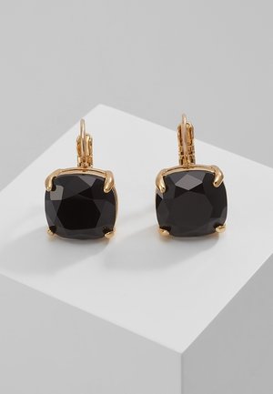 Złote kolczyki z dużymi czarnymi kamieniami szlachetnymi w kształcie poduszki, osadzone na czterech łapkach, wystawione na białej geometrycznej platformie.