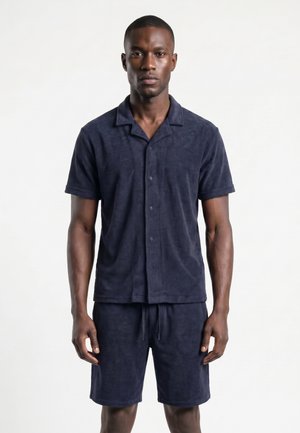 Homme debout face à l’objectif portant un ensemble chemise boutonnée à manches courtes et short bleu foncé assortis sur un fond uni.