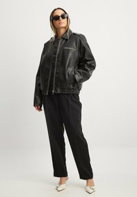 Jacca bomber in pelle nera con chiusura zip, tasche laterali e orlo a coste; abbinata a pantaloni neri e scarpe slip-on bianche.