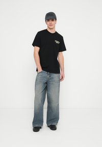 T-shirt preta de algodão com um pequeno logótipo amarelo no peito esquerdo; combinada com jeans de ganga azul clara e de corte largo. Sapatos pretos e uma boné escura.