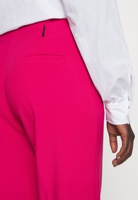 Fuchsia op maat gemaakte broeken met een achterzakdetail, gecombineerd met een witte blouse met lange mouwen en een manchet detail. Glad textuur.
