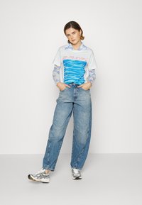 T-shirt branca com gráfico de onda azul e texto vermelho, combinada com jeans de perna larga azul claro. Ténis prateados e pretos completam o visual.