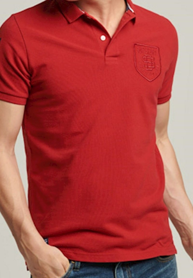 Homme portant un polo rouge à manches courtes avec un petit blason brodé sur le côté gauche de la poitrine, assorti à un jean bleu.