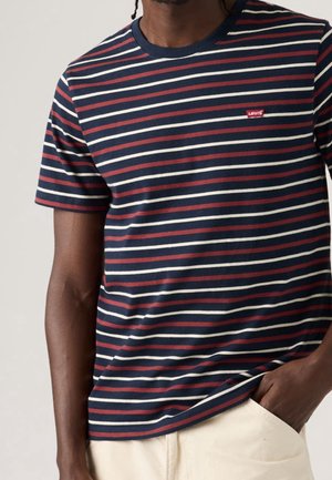 Mand iført en marineblå, rød og hvid stribet Levi's t-shirt med korte ærmer og beige bukser, hånd i lommen.
