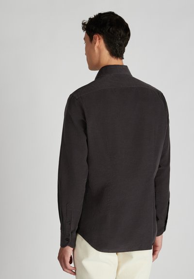 Man in een donkergrijs overhemd met lange mouwen en lichte broek, weergegeven van achteren tegen een effen grijze achtergrond.