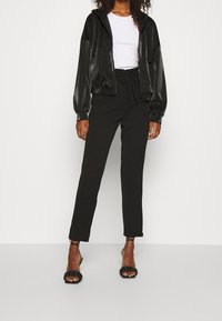 Zwarte, glanzende zip-up jack met een capuchon, gecombineerd met aansluitende zwarte broeken en zwarte sandaaltjes met bandjes. De outfit heeft een witte T-shirt eronder.