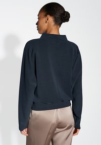 Pullover navy con maniche lunghe e colletto a imbottitura; presenta un orlo a coste e una texture di tessuto liscia. Indossato con pantaloni satin beige.