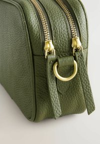 Groene leren cosmetica tas met een gestructureerde oppervlakte, dubbele gouden ritsen, draagriem met een gouden ringaccent en een rechthoekige vorm.