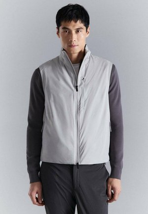 Hombre con chaleco sin mangas gris claro con cremallera sobre camisa de manga larga oscura y pantalones oscuros, de pie contra un fondo gris liso.