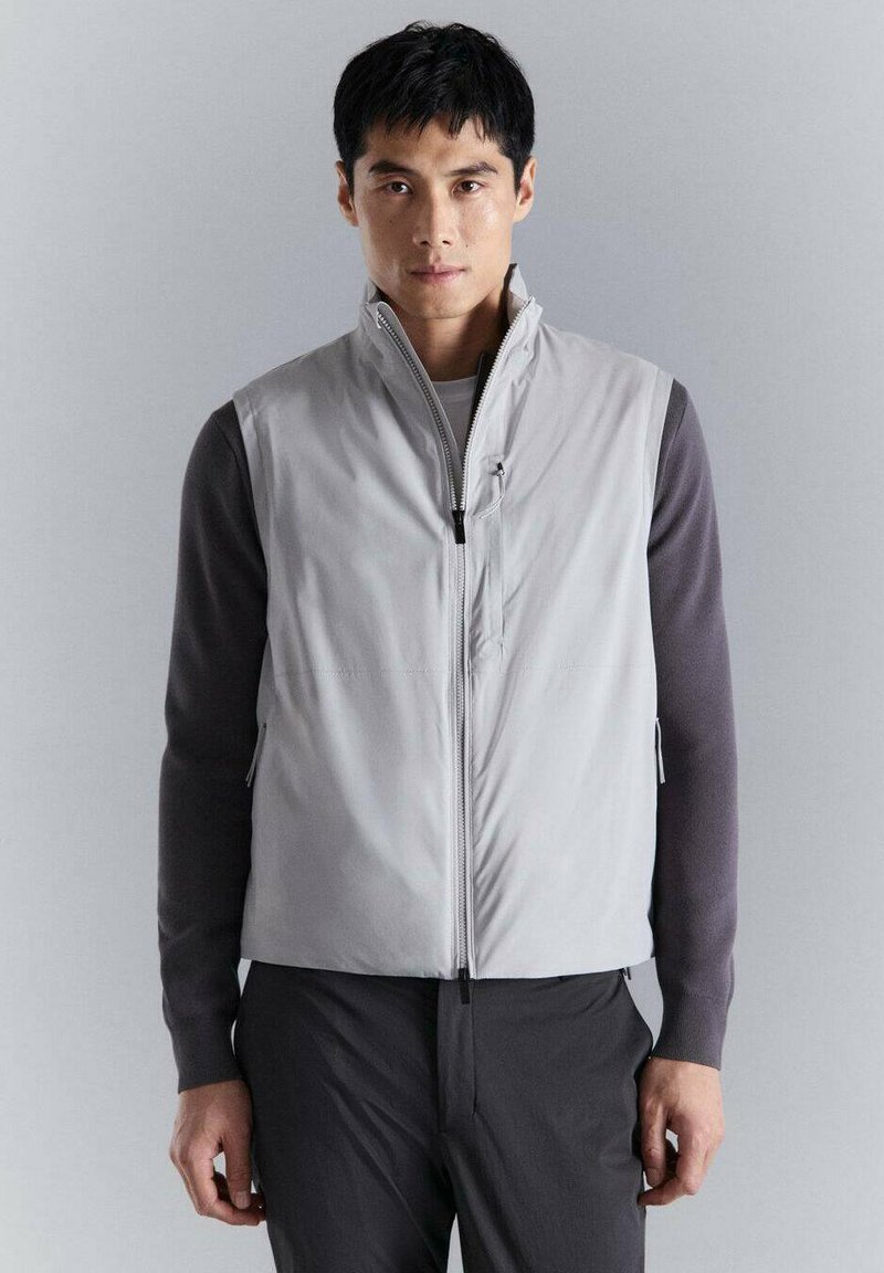 Uomo che indossa un gilet grigio chiaro senza maniche con zip sopra una camicia scura a maniche lunghe e pantaloni scuri, in piedi contro uno sfondo grigio uniforme.