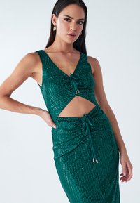 Žalia, trikampės tekstūros su akutėmis suknelė su v formos iškirpte, išpjova ir raišteliais juosmens srityje. Lygi tekstūra ir lieknos formos dizainas.