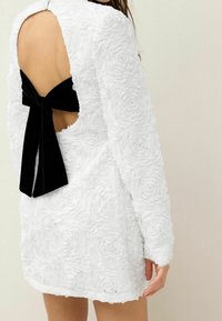 Robe blanche avec des appliqués de roses texturés, manches longues, dos ouvert et détail en velours noir en nœud à la taille.