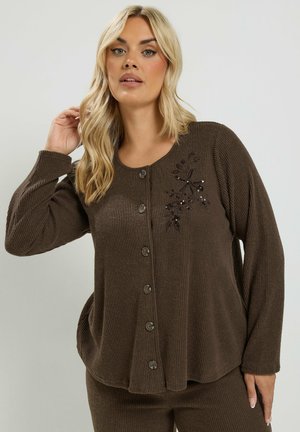 Donna con capelli biondi che indossa un cardigan marrone a coste con bottoni, ricamo floreale sul petto e pantaloni coordinati.