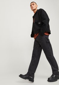Zwarte denimjas met een borstzak, gecombineerd met een roestkleurig T-shirt en zwarte cargo-broeken. Stevige zwarte boots maken de look compleet.