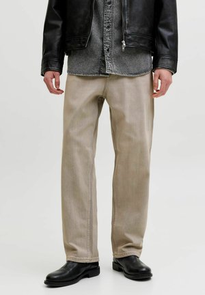 Mann trägt beige gerade geschnittene Hose, schwarze Chelsea-Lederstiefel, Jeanshemd und schwarze Lederjacke, steht auf schlichtem hellem Hintergrund.