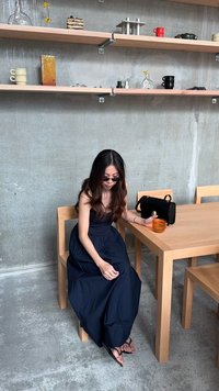 O masă din lemn cu un finisaj deschis, însoțită de o persoană îmbrăcată într-o rochie de un navy închis. Aceștia țin o băutură portocalie. O geantă neagră stă alături.