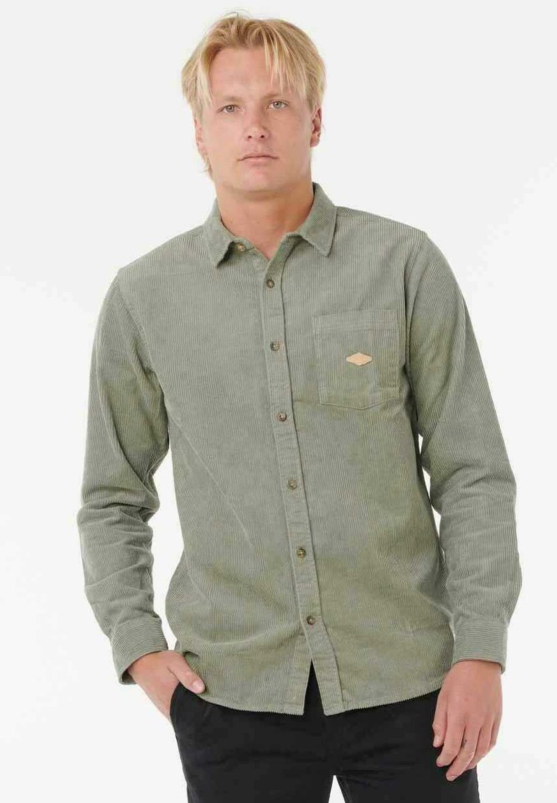 Camisa de pana verde con cuello abotonado, mangas largas, un bolsillo en el pecho y un acento marrón claro cerca del bolsillo. Tela texturizada.