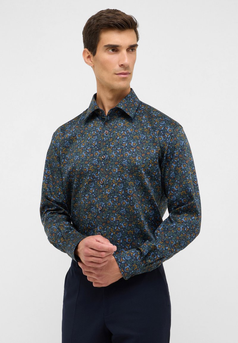 Uomo che indossa una camicia a bottoni scura con un motivo floreale, caratterizzata da fiori blu e arancioni, in piedi con le mani incrociate, mentre guarda leggermente verso destra.
