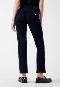 Jeans di denim neri con silhouette aderente, due tasche posteriori e un piccolo logo triangolare sulla cintura. Abbinati a mocassini neri.