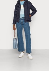 Person mit einer marineblauen Steppjacke, gestreiftem blau-weißen Pullover, verkürzten blauen Jeans, weißen Sneakers und einer hellblauen Handtasche.