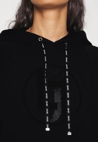 Primer plano de una sudadera negra con un logo circular brillante y cordones negros impresos con el texto blanco "LIUJO", llevada por una persona con cabello largo y oscuro.