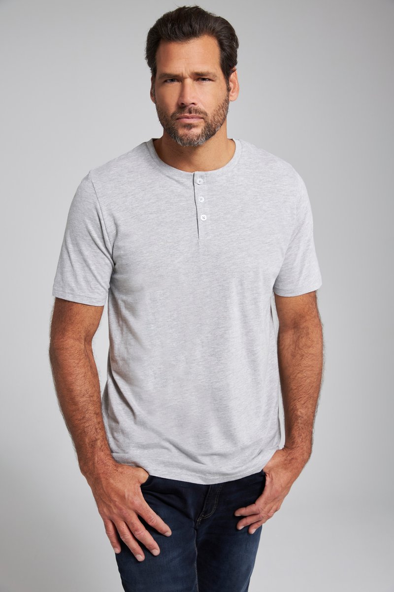 Man met donker haar en baard, dragen een lichtgrijs henleyshirt met korte mouwen en een donkere spijkerbroek, staande voor een effen grijze achtergrond.
