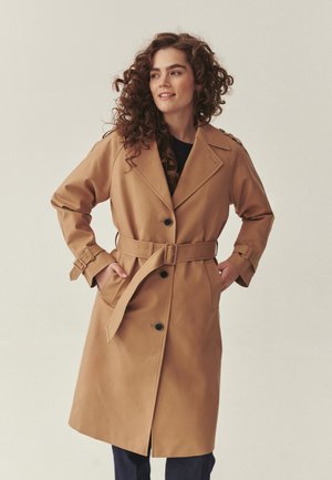 Kvinde med krøllet hår iført en beige trenchcoat med bælte og hænderne i lommerne, der kigger til siden mod en ensfarvet baggrund.