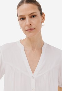 Camicia bianca a maniche corte, con bottoni, taglio dritto e scollatura a V che presenta un delicato dettaglio di pieghe lungo il busto. Tessuto morbido.