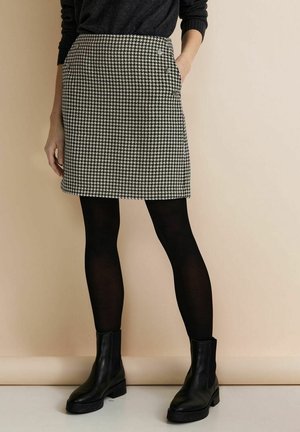 Person trägt einen schwarz-weißen Hahnentrittrock, schwarze Strumpfhosen, schwarze Stiefel und ein schwarzes langärmeliges Oberteil und steht vor einem beigen Hintergrund.