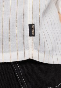 Tejido blanco con finas rayas verticales doradas y una etiqueta negra "Superdry" cosida cerca del dobladillo, encima de una tela de mezclilla negra cosida.