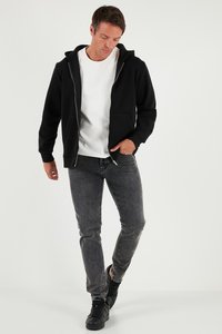 Crna zip-hoodica s prednjim džepovima, slojevita preko bijele majice, u kombinaciji s tamno sivim trapericama i crnim tenisicama. Glatka tekstura tkanine.