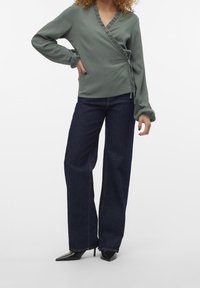 Grön texturerad omlottopp med volangkrage, ihop parat med mörkblå vida jeans och spetsiga svarta klackar. Avslappnad, lager-på-lager outfit.